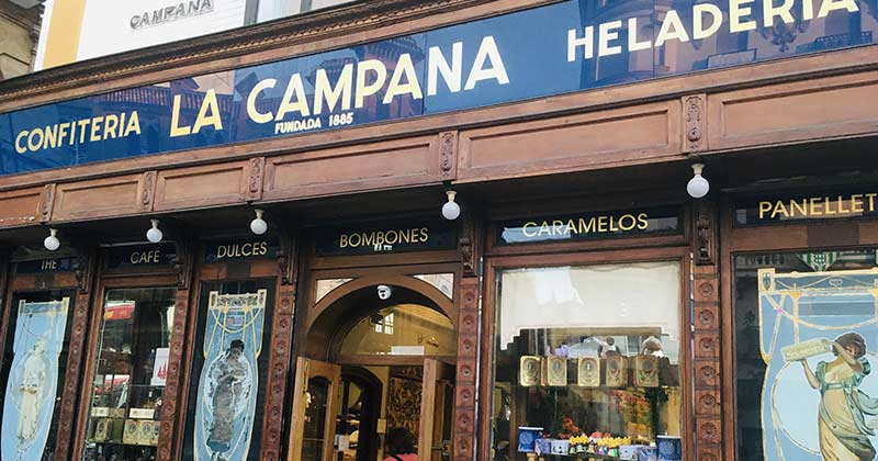 La Campana in Seville features blue and gold signs, wooden details, and window displays of sweets and colorful posters.