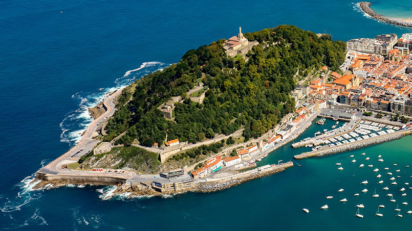 A lush green hill bordered by a winding road sits by turquoise water, marina with boats, and a city of red-roofed buildings.