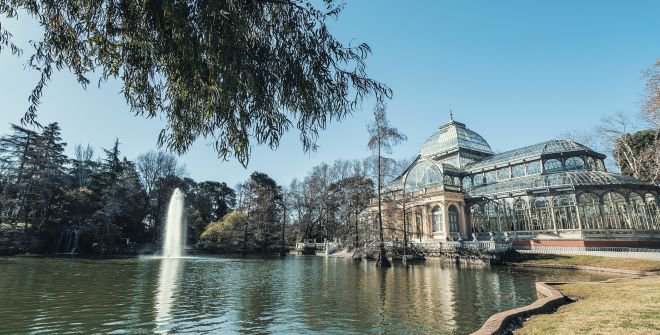 The Glass Palace at Madrid’s Retiro Park reflects in a pond, fountain spraying; tree branches frame the view under blue sky.