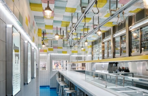 People sit at stainless steel bar stools in a well-lit Madrid diner with pastel ceiling tiles, posters, and bottles on the walls.