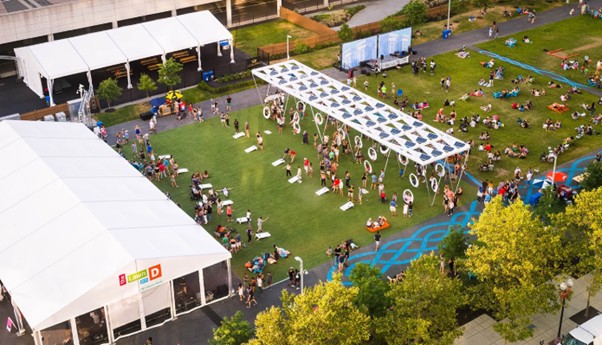 People gather in a Boston urban park in spring, relaxing on grass, sitting under tents, and socializing in small groups.