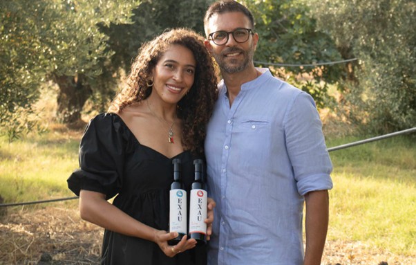 A woman in a black dress holds two olive oil bottles beside a smiling man in glasses; they stand together among leafy olive trees. This EXAU Olive oil is an ideal gift for travel lovers.