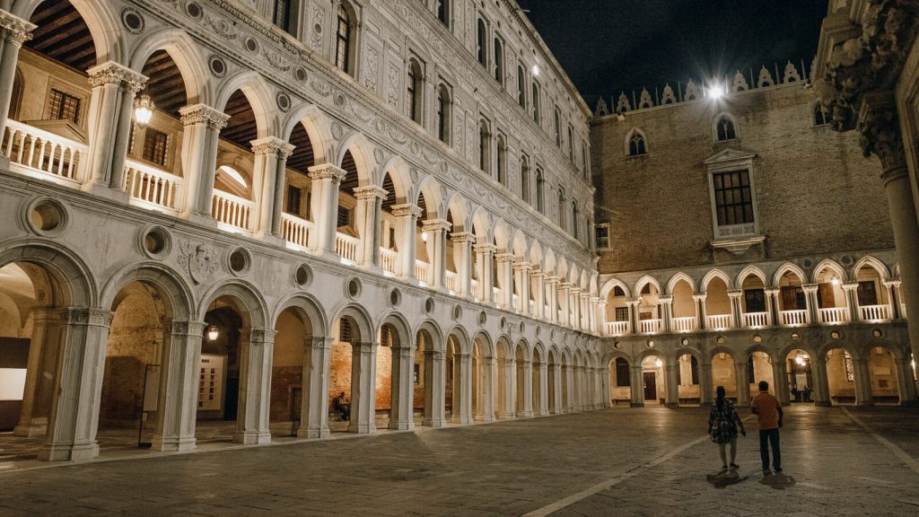 At night, two people walk beneath glowing arches and columns in a grand courtyard lined with balconies and tall windows.