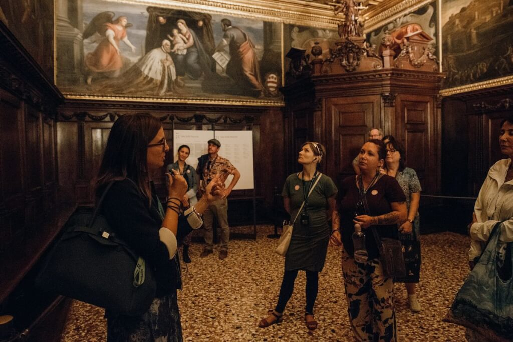 A tour guide addresses visitors in a grand wood-paneled gallery with gold-trimmed ceilings and artwork lining the walls.