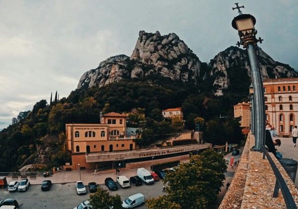 Montserrat mountain rises under cloudy skies, with buildings and parked cars below, viewed from a stone terrace with a lamp post.