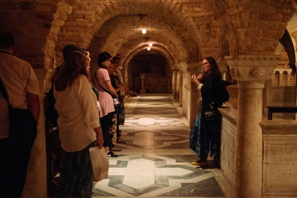 Several visitors listen to a guide in a dark, arched stone hallway with patterned tile floors, likely inside a historic building.
