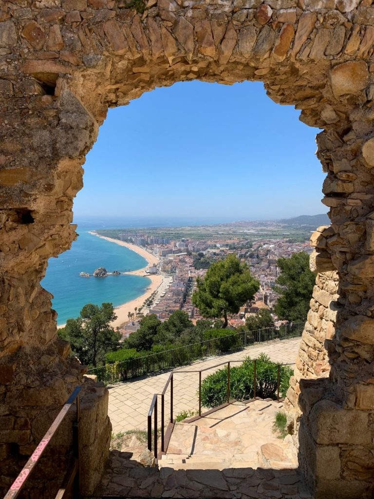Looking through a stone archway in ancient ruins, steps descend toward a sandy beach town near barcelona and the blue Mediterranean Sea.