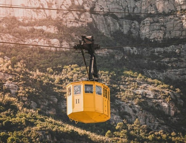 A yellow cable car marked 11 moves along cables over Montserrat, Spain, above rocky cliffs and dense green hills.