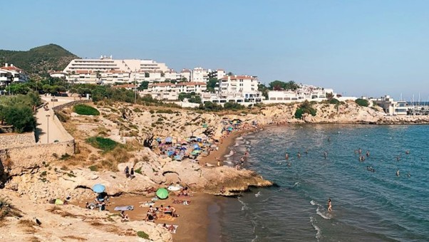 People lounge under umbrellas on a beach near Barcelona as swimmers enjoy calm water; white buildings and green hills behind.