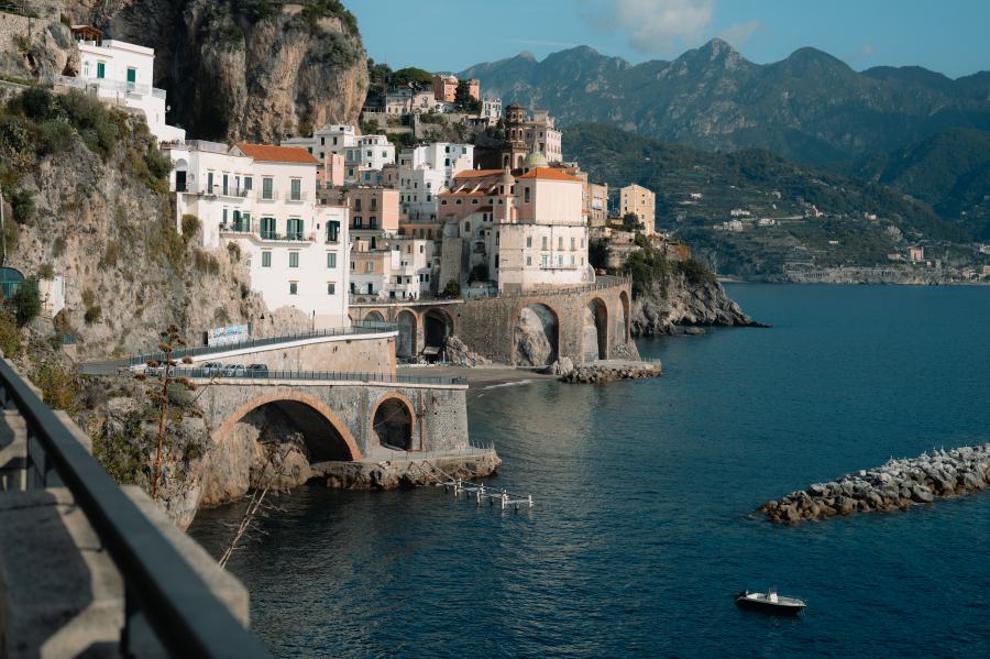 A small boat approaches pastel houses on steep cliffs in Amalfi, Italy, with arched bridges and green mountains behind.
