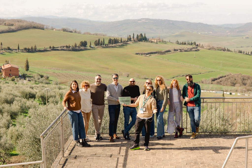 Ten people smile as they enjoy a multi day tour from Rome, in the tuscan countryside with green hills, trees, and a farmhouse behind them under clouds.