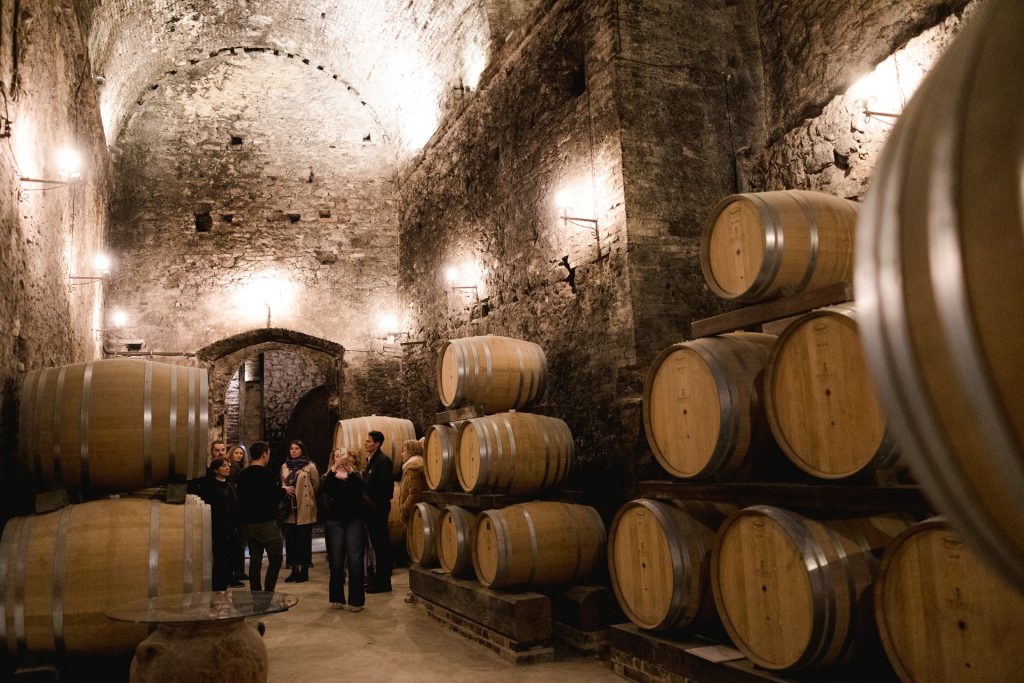 Several people walk through a dimly lit stone wine cellar near Rome, surrounded by large wooden barrels and arched ceilings.