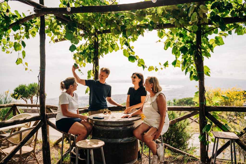 Four people chat and laugh around a wooden barrel table under a leafy pergola, surrounded by green hills near Rome.