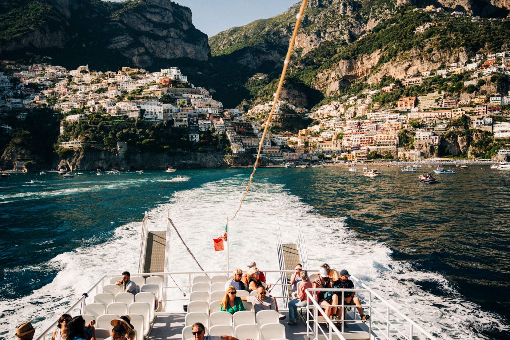 Several people relax on a boat deck, departing a colorful Italian coastal town with hillside houses and deep blue water, this experience can be enjoyed as part of an overnight trip from Rome.