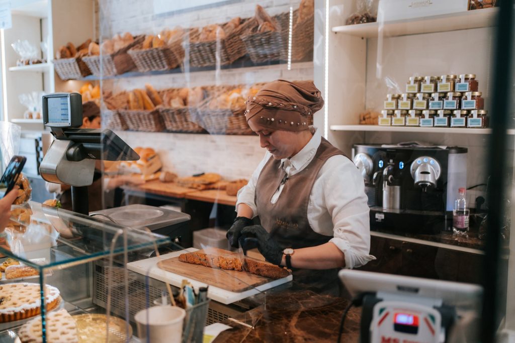 A bakery worker in a brown headscarf and apron slices bread behind a counter lined with pastries and jars in an Italian bakery.