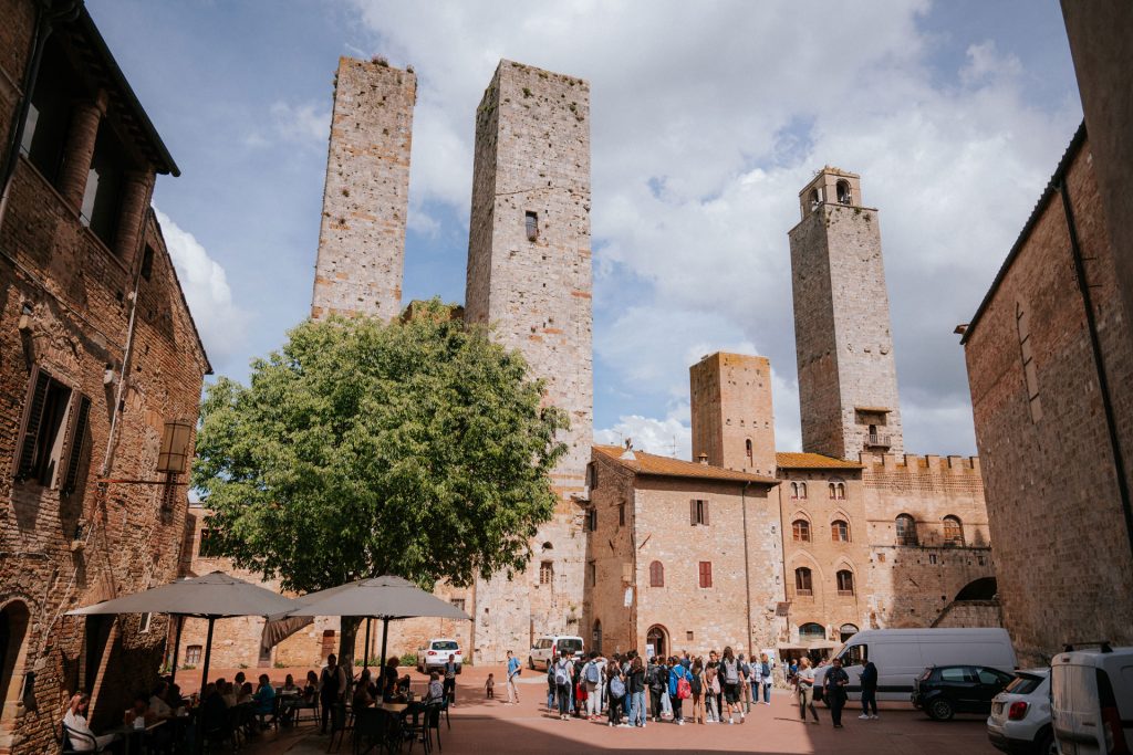 Tourists stand in San Gimignano’s sunny square, surrounded by medieval towers, as people relax at a café under umbrellas.
