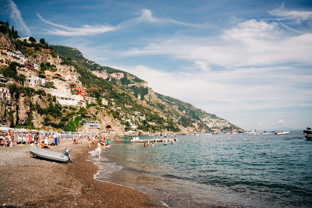 People swim and sunbathe on a lively beach by rocky cliffs and colorful buildings, boats passing under a bright blue sky on the Amalfi Coast, just an overnight trip from Rome.