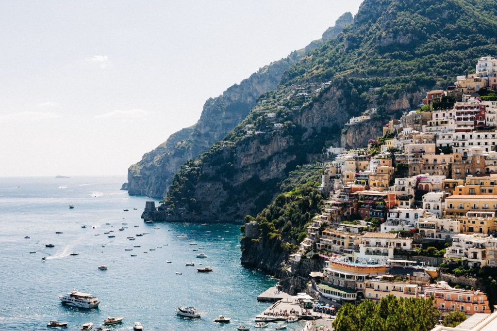 Brightly painted houses cascade down a coastal hillside in Positano, Italy, with boats scattered on the blue sea and lush cliffs behind. This is the Amalfi Coast, an ideal place to visit on an overnight trip from Rome