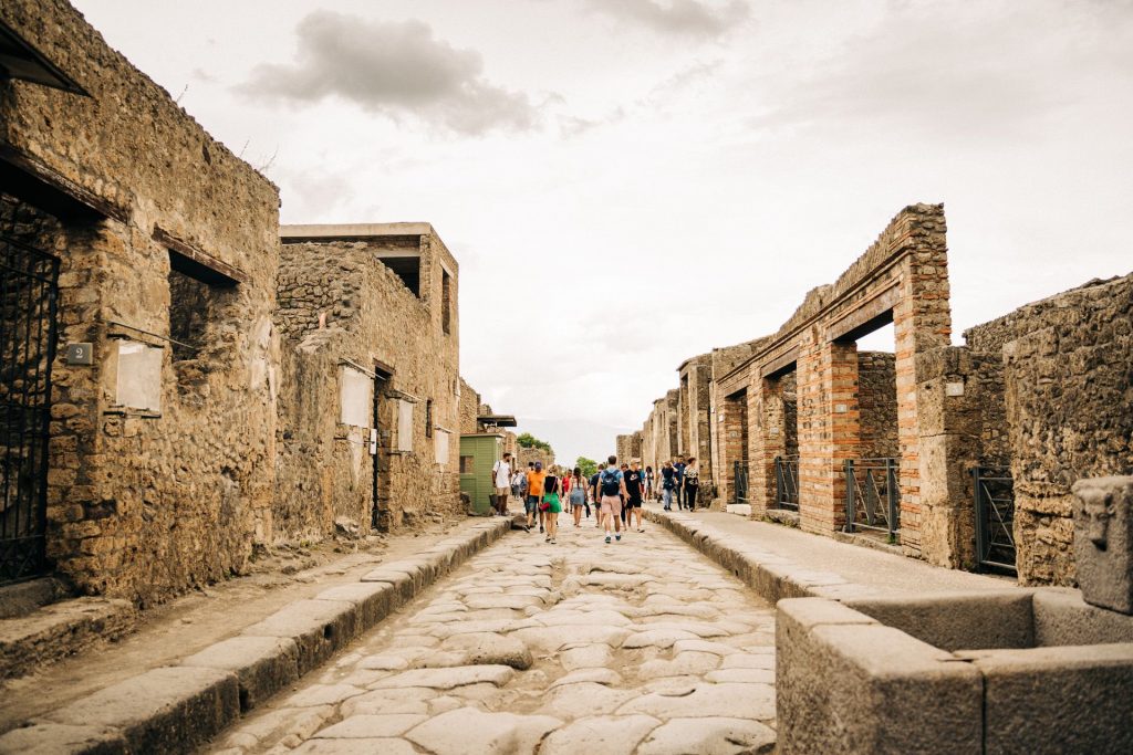Tourists stroll down a broad, ancient stone street in Pompeii, passing ruined buildings beneath a cloudy sky.