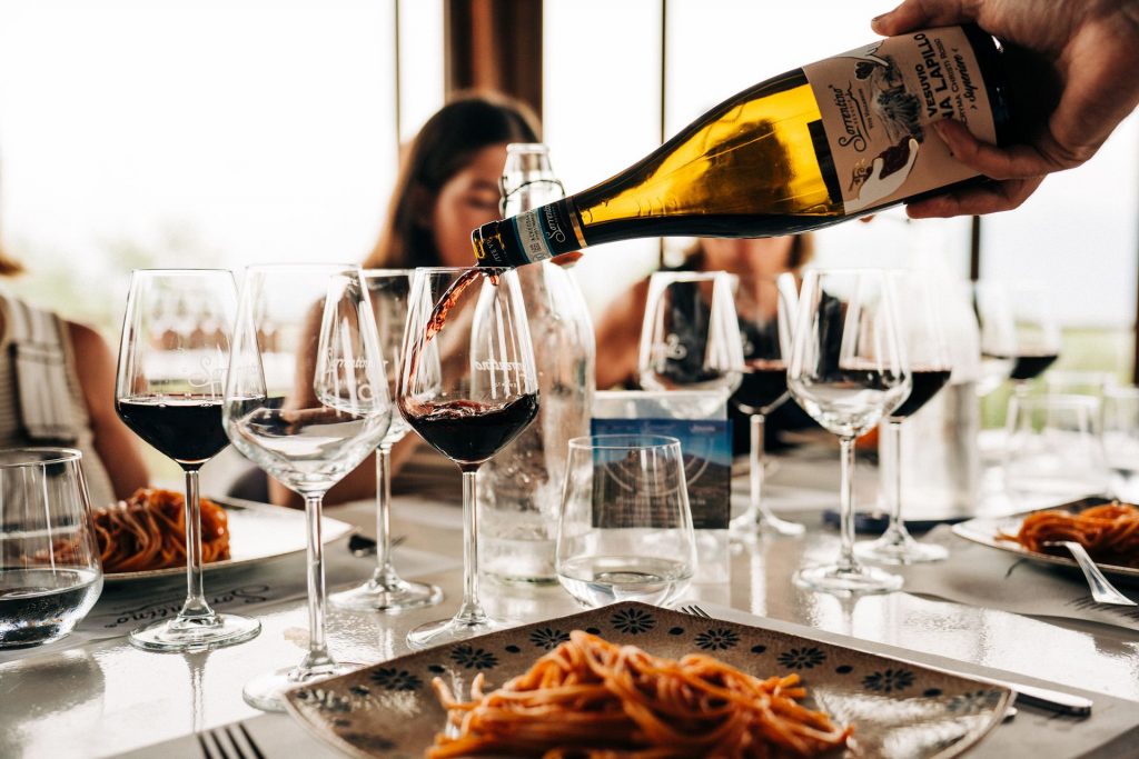A hand pours red wine into a glass at a table with spaghetti and drinks, while people dine together on an overnight trip from Rome.