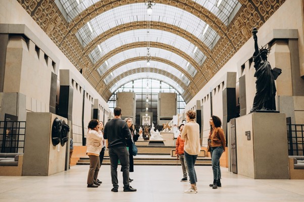 People converse in the Impressionist Museum, Paris, under arched ceilings near sculptures and a small Statue of Liberty replica.