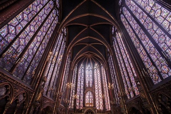 Sunlight streams through tall, ornate stained glass windows in a gothic cathedral, coloring columns and arches with vibrant patterns.