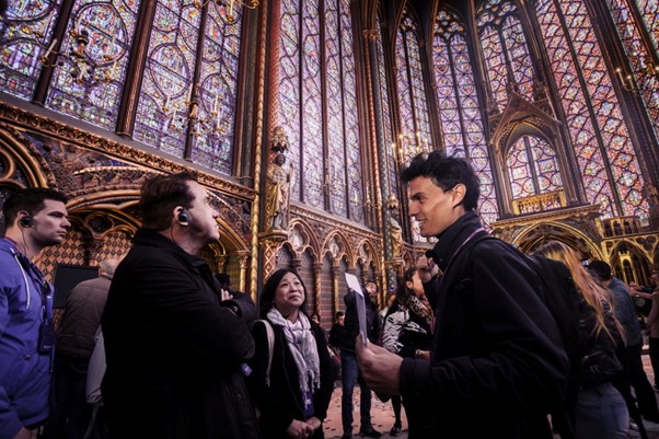 Tourists wearing audio guides listen attentively as they admire Sainte Chapelle’s stained glass windows and ornate gold detailing in Paris.