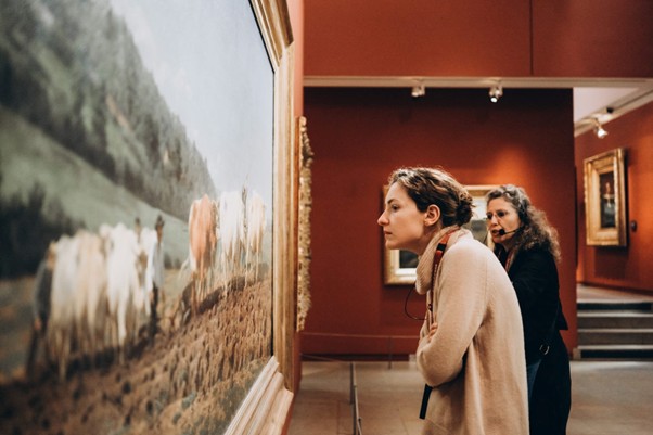 Two women study Rosa Bonheur’s “The Cows” at the Impressionist Museum in Paris, surrounded by red walls and framed art.