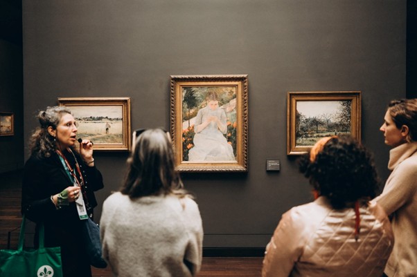 A guide at the Impressionist Museum in Paris discusses Monet’s “Woman in White” with three visitors before gray gallery walls.