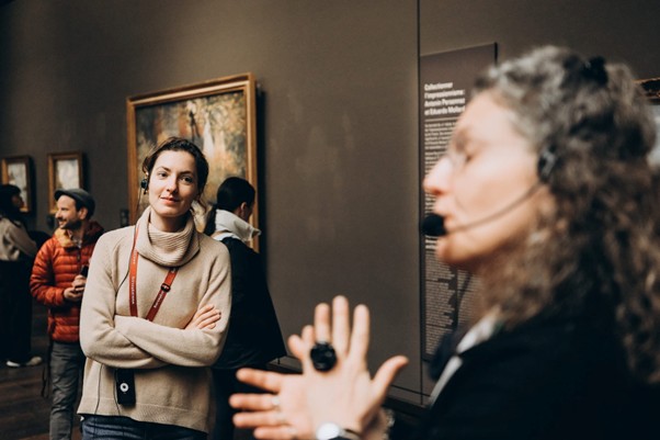 At the Impressionist Museum in Paris, a woman with crossed arms listens to a guide; blurred Monet paintings hang behind her.