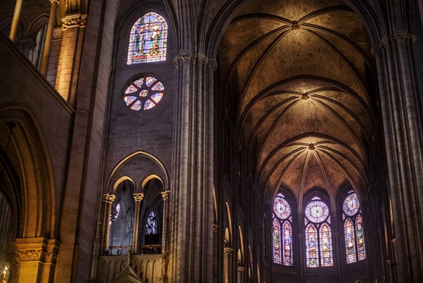 Inside Sainte Chapelle, sunlight shines through ornate stained glass windows, casting colorful patterns on the Gothic arches and vaulted ceiling.