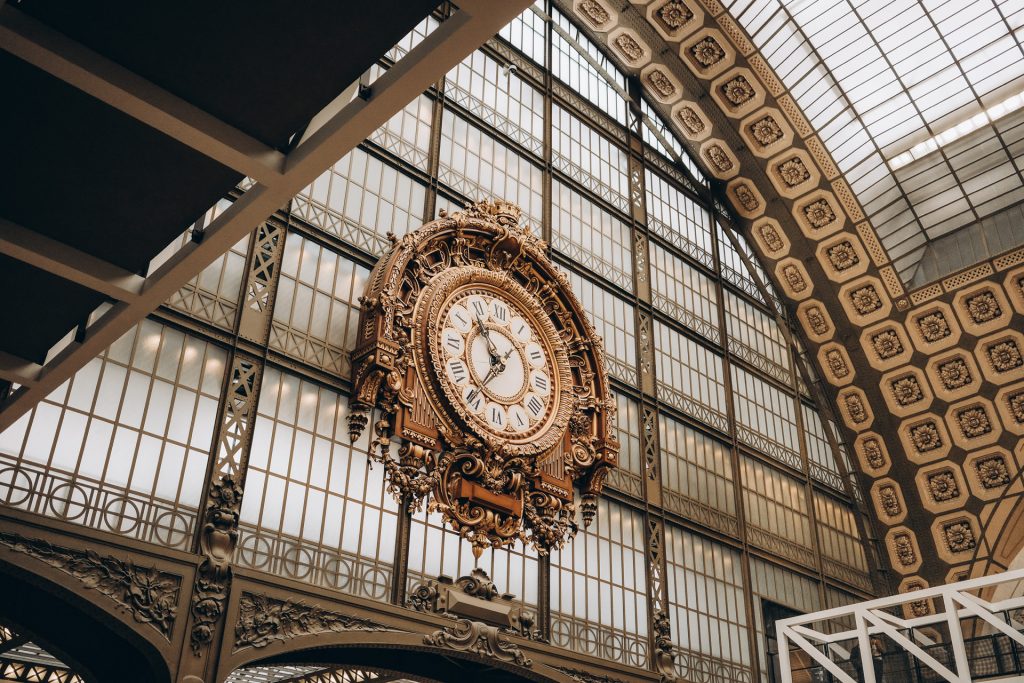 An ornate gold clock hangs on a decorative wall in Musée d’Orsay, Paris, beneath an arched glass and metal ceiling.